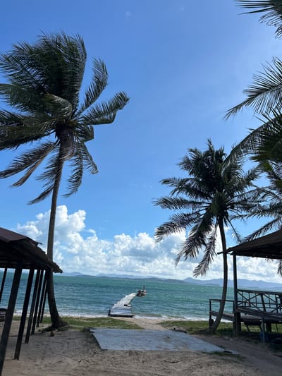 The image depicts a serene tropical beach scene in Ban Khlong Hia, Thailand. The foreground is dominated by a sandy beach with two open-air structures, one on the left and another on the right, providing shade. Palm trees with lush green fronds are scattered across the scene, with two prominent ones framing the view of the ocean. The ocean itself is a vibrant turquoise, with moderate waves lapping at the shore. A white wooden pier extends into the water, leading towards a distant boat. In the background, a hazy silhouette of islands or mountains can be seen against a bright blue sky dotted with fluffy white clouds. The overall impression is one of a peaceful and idyllic coastal getaway.