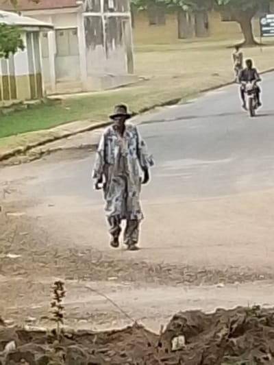 The image shows an outdoor scene, likely on a paved or dirt road, under what appears to be daylight conditions. The central and most prominent figure is a person walking towards the viewer, slightly off-center to the left. This individual is wearing a dark, wide-brimmed hat, possibly a fedora. Their main attire consists of a long, loose-fitting garment, resembling a robe or coat, with a mottled or patterned design in shades of grey, white, and possibly light blue or green. Underneath this garment, trousers are visible, and the person is wearing sandals or open-toed shoes.

In the mid-ground, to the right of the central figure, another person is seen riding a motorcycle away from the viewer. Further down the road and to the right, a third person is walking. The road appears to have a slight curve or incline.

The background features several elements:
- To the far left, there is a building with light-colored walls and what looks like a dark stripe or trim. Green foliage is visible next to and above this building.
- Behind the central figure and to the left, there's a taller, light-colored structure that appears somewhat weathered, possibly a tower or a part of another building.
- Towards the right and further back, there are trees with green leaves and hints of other structures. The ground to the sides of the road is grassy or covered in sparse vegetation.

The foreground is blurry and consists of an earthy, reddish-brown ground with some indistinct plant life. The overall image quality is somewhat soft and slightly out of focus, giving it a painterly or impressionistic feel. There is no visible text in the image. The setting suggests a casual, everyday street scene in a potentially rural or semi-urban environment.
