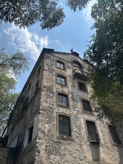 The image depicts a tall, old stone building with multiple windows, some with bars, framed by trees and the sky. The building appears to be deteriorating, with patches of discoloration and missing plaster. There are hints of brickwork beneath the stone facade. The image is taken from a low angle, looking up. Based on its architectural style and visible features, it is likely located in Tlalnepantla, Mexico, given its mention. The windows are rectangular, except for one arched window at the top. There is a small balcony or ledge near the top of the building. The sky is blue with scattered clouds. No people are visible.