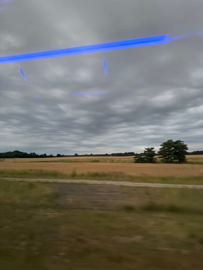 The image depicts a rural landscape under a cloudy sky, taken from a moving vehicle, likely a train or bus, given the motion blur and the reflection of blue LED lights on the window. The scene is characterized by a wide expanse of golden, harvested fields, suggesting the summer or early autumn season. A line of dark green trees borders the fields in the distance, with a few scattered trees closer to the foreground. The sky is filled with heavy, gray clouds, creating a subdued and atmospheric lighting. In the lower portion of the frame, a blurred strip of green vegetation and a gray horizontal line, possibly a road or a path, are visible. There are no people or animals visible in the image, and the overall impression is one of quiet, natural scenery. There is no indication of the specific location within Mozów, Poland, other than it being a rural area.