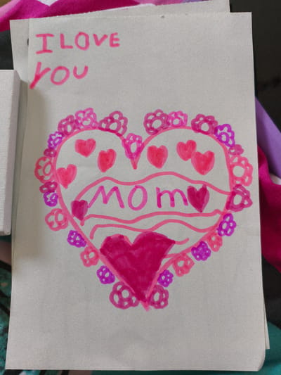 The image shows a handmade card with a drawing of a heart. The text "I LOVE YOU" is written in pink marker at the top of the card. Inside the heart, the word "MOM" is written in larger pink letters, with wavy lines underneath. The heart is outlined with pink and purple flowers, and there are smaller pink hearts scattered inside the main heart. The card is on a white surface, possibly a table, with a hint of colorful fabric in the background. There are no people or animals visible in the image, and no specific details are present that would indicate a location in Ramla, Israel.