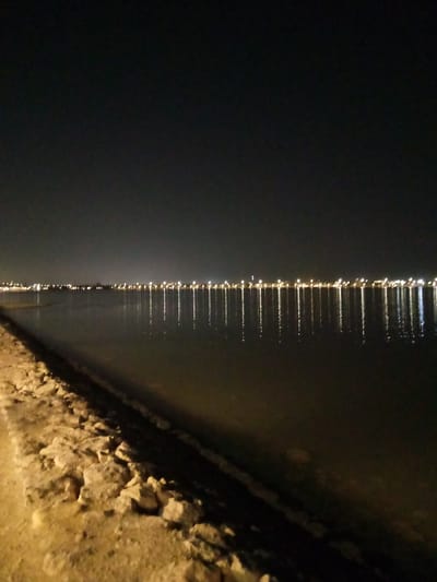 This media file captures a serene night scene in Al Khobar, Saudi Arabia, focusing on a body of water bordered by a shoreline. The dominant feature is a long, continuous line of bright artificial lights stretching across the distant horizon, likely from a city or settlement. These lights are clearly reflected as vertical streaks of varying intensity on the calm water surface, creating a visually striking pattern.
In the foreground, along the left side of the frame, there is a rough, rocky, and sandy bank or shore. The ground here is illuminated by an unseen light source, casting a warm, yellowish glow on the terrain and highlighting the individual rocks and textured surface. The water itself appears dark and relatively still, allowing for crisp reflections. The sky above is a deep, uniform black, indicative of night.
There are no people or explicit activities visible in the image, emphasizing the quiet solitude of the nocturnal landscape. The calm water surface and clear reflections suggest tranquil weather conditions without significant wind or disturbance. The overall impression is one of peaceful urban beauty at night, with the distant city lights contrasting against the dark expanse of the water and sky. No visible text is present in the image.