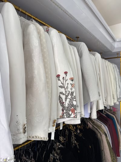 The image displays a row of white jackets hanging on a golden rack. The jackets are varied in texture and embellishment, with some featuring intricate embroidery of floral designs, beadwork, and metallic threads. The lighting in the scene is bright, suggesting an indoor retail or studio environment. The background is simple and uncluttered, with a white ceiling that appears to have integrated LED lighting strips. Below the row of white jackets, a second rack is visible, holding darker colored garments. The overall impression is one of a curated collection of formal or ceremonial wear. The specific location in Tanjungmeru, Indonesia, cannot be determined from the visual information provided.