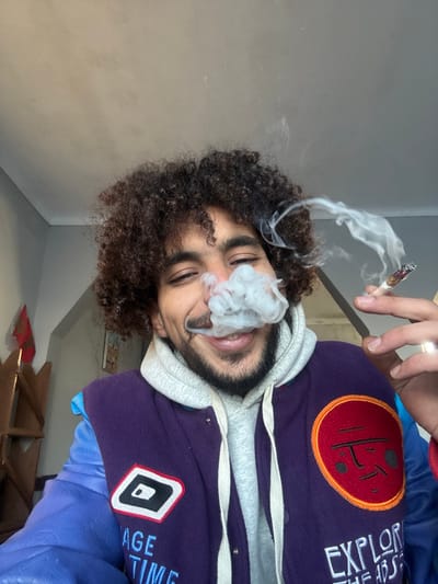 A person with curly hair and a beard holds a lit cigarette, exhaling smoke. They are wearing a purple jacket with patches and a gray hoodie. The jacket has patches that read "EXPLORE" and "VINTAGE TIME". They are smiling, and a ring is visible on one of their fingers. The background shows a corner of a room with light-colored walls and a wooden shelf.
