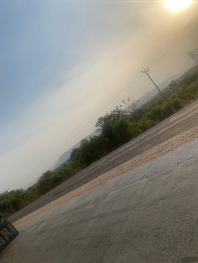 The image captures an outdoor scene, appearing to be a rural or semi-rural road alongside a natural landscape, seen during what looks like late afternoon or early morning due to the low sun and hazy atmosphere. The image is oriented diagonally, with the horizon line running from the top left to the bottom right if viewed correctly.
In the upper left quadrant, a bright, intense sun is visible, creating a strong lens flare effect and casting a warm, golden glow across the sky. The sky itself transitions from a bright, hazy yellowish-white near the sun to a pale blue-grey further away, suggesting a significant amount of atmospheric haze or fog.
A concrete or asphalt road occupies a significant portion of the right side of the image, running diagonally from the top right down towards the bottom left. The surface of the road appears textured and dry. A strip of lighter-colored, possibly sandy or gravelly ground runs along the edge of the road, separating it from the natural landscape.
To the left of the road, a line of dense green trees and vegetation stretches across the midground. Beyond these trees, a range of mountains or hills is faintly visible in the far background, obscured by the atmospheric haze.
Several utility poles and power lines are visible, extending across the scene. One prominent pole stands roughly in the center-left, with multiple lines stretching horizontally across the sky and above the treeline. Some leafless or sparse trees are also scattered among the denser green foliage.
In the very bottom right corner of the image, a small portion of what appears to be a colorful object or vehicle is visible, possibly a motorcycle or a cart, with some indistinct shapes and colors, and what might be the letters "NR" or "RN" in a stylized font, though this is difficult to discern clearly due to the crop and angle.
The overall impression is one of a quiet, somewhat hazy day in a natural setting, with human infrastructure (road, power lines) integrated into the landscape.