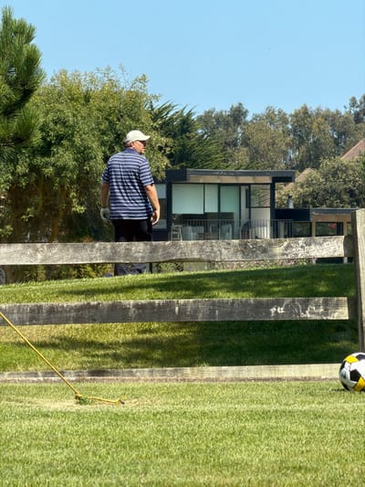 This image depicts an outdoor scene in Santo Domingo, Chile, under clear blue skies with bright sunlight. In the foreground, a well-maintained grassy lawn is visible. A yellow rope or cord lies diagonally across the grass in the bottom-left corner, with a knot visible near its end. A black, white, and yellow soccer ball is partially visible in the bottom-right corner of the grass.
A weathered wooden fence, consisting of two horizontal rails and vertical posts, spans the mid-ground, acting as a barrier. Beyond this fence, to the left of the frame's center, stands a male individual, appearing middle-aged. He wears a light-colored baseball cap, a short-sleeved polo shirt with horizontal navy blue and white stripes, dark trousers, and a white glove on his left hand. He is oriented facing right, with his back partially towards the viewer, appearing to be in a paused or walking stance.
Behind the man and the fence, a modern-style residential building is visible, featuring a flat dark roof, large glass windows (some with white blinds), and a balcony with a light-colored railing. The building is partially obscured by a dense cluster of various green trees and shrubs, including tall conifers and leafy deciduous trees, which dominate the background. The upper portion of the image is filled with a clear, bright blue sky.