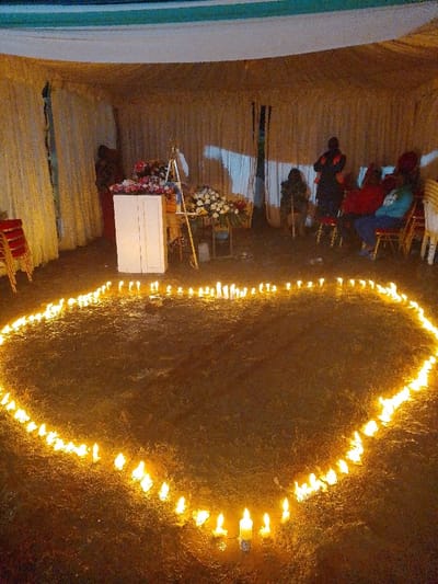 The image depicts an organized event space within a temporary structure, likely a tent, situated in Marenyo-Shianda ward, Kenya. The environment is dimly lit by artificial sources and numerous candles, suggesting a nighttime or low-light setting. The tent features cream-colored fabric walls and a ceiling with sections of white and dark blue/green material.
In the foreground, a large heart shape is meticulously outlined on the dark, earthen or sparsely vegetated ground by approximately 70-80 individual lit white candles. The flames cast a warm glow across the immediate area.
In the background, positioned against the rear tent wall, a white, two-door cabinet or podium serves as a central point, adorned with multiple bouquets of red and white flowers. To its right, a smaller, ornate metal stand also holds floral arrangements, and an easel-like structure is visible behind the cabinet. A stack of red chairs is discernible on the far left.
To the right side of the background, approximately five individuals are present. One figure stands in dark clothing with a red accent, while others are seated on red and white chairs, appearing to observe the scene. Their attire is varied, including light blue and red tops. No direct interactions between individuals are clearly depicted.