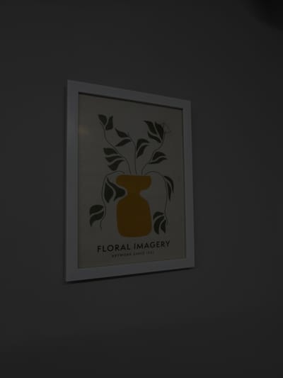 The image shows a framed print hanging on a wall. The print depicts a stylized floral arrangement with a yellow vase and dark green leaves. The text "FLORAL IMAGERY" and "ARTWORK SINCE 1921" is printed on the right side of the artwork. The frame is white. The background is a plain, dark gray wall. The location is not discernible from the image. There are no people or interactions visible.