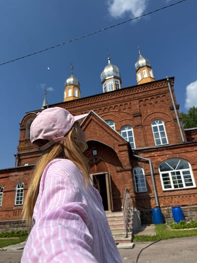 A young woman, seen from the back, stands on a paved area in front of a brick building with an arched entrance and multiple domes, likely a church, under a clear blue sky. She is wearing a pink and white striped shirt and a pink baseball cap. The building exhibits architectural details such as arched windows and decorative brickwork. Two large blue barrels are visible near the base of the building. The scene suggests an outdoor, possibly historical, location.