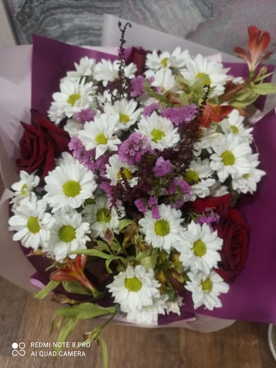 A top-down medium shot captures a bouquet of flowers against a textured, grey background and a wooden floor. The bouquet is artfully arranged with white daisies, deep red roses, delicate purple flowers, and a few orange lilies. The flowers are wrapped in alternating layers of purple and pale pink paper. The overall impression is one of natural beauty and thoughtful presentation. The image was taken in Kugesi, Russia.