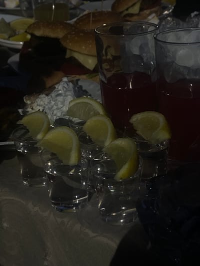 The media file depicts a tabletop scene, likely indoors given the lighting, with a focus on beverages and food. The dominant objects are several clear glasses, some filled with ice, and others appearing to be empty or holding a dark red liquid. Wedges of lemon are placed on the rims of some of the glasses, suggesting they are being prepared for drinks.

In the background, blurry elements suggest a dining or party setting. There are glimpses of what might be food items, possibly burgers or sandwiches, and a large container of red liquid. The lighting is dim and appears artificial, creating strong shadows and highlights, which makes it difficult to discern specific details of the background or exact time of day. The overall impression is that of a gathering or a preparation for a meal or event.

The image is oriented in a way that suggests it might have been taken at an angle or that the objects themselves are arranged on a tilted surface. No people are clearly visible, though their presence is implied by the context of prepared food and drinks. No text is discernible in the image. The location context provided is Mala tsarkva, Bulgaria, but there are no visual cues in the image itself that directly confirm this specific location.