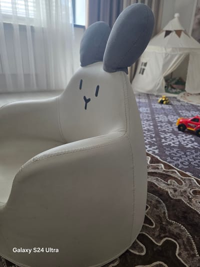 A white, plush children's armchair shaped like a cartoon rabbit with grey ears is positioned in the foreground, facing right. The rabbit's face is depicted with simple black lines for eyes and a mouth. In the background, a white play tent with a conical roof and windows is visible. A patterned rug covers the floor. Toy vehicles, a red truck and a yellow car, are placed on the rug near the tent. Sheer white curtains hang in front of a window on the left side of the image. The overall setting appears to be a child's play area, likely indoors, with soft lighting. There are no people in the image. The location is not specified in the image.