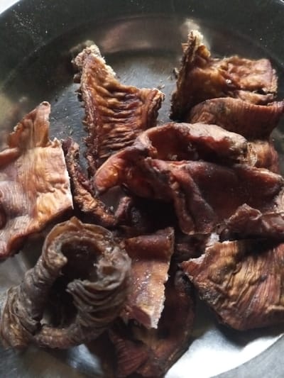 In a metal bowl, there's a pile of cooked or smoked organ meat, potentially intestines. Some pieces are curled, displaying visible ridges or folds. The color ranges from light brown to a darker, reddish-brown. The meat appears slightly dry and crispy in texture, with a few bits of white or light-colored areas possibly being fat or connective tissue. The pieces are irregularly shaped and cut into varying sizes.