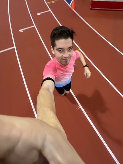 A high-angle selfie captures a person standing on an indoor running track. The camera is held by the person's outstretched right arm, pointing downwards, creating a slightly distorted perspective where the person's head appears larger and their body tapers towards their feet.
The person is a male with dark hair, looking up at the camera with a slight smile. He is wearing a short-sleeved athletic t-shirt that transitions from a vibrant pink at the top to a light blue at the bottom. He has black shorts on and black athletic shoes. On his left wrist, he wears a black fitness tracker or watch, and a white earbud is visible in his left ear. Another black wristband is on his right wrist, which is holding the camera.
The setting is a reddish-brown synthetic running track, marked with crisp white lane lines. The lines are visible across the frame, indicating multiple lanes. The track appears to be indoors, suggested by the uniform lighting and what looks like a wall or barrier on the far left edge of the frame, near an orange cone. The person's shadow is cast prominently on the track behind and to their right, elongated due to the overhead light source. No text is visible in the image. The overall impression is one of a brief pause during an athletic activity, possibly before or after a run or workout.