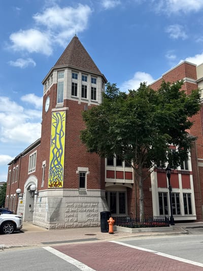 This image depicts the exterior of a building located at 139 S Main St, Naperville, IL 60540, United States of America. The building is constructed with red brick and features a prominent clock tower with a shingled roof. A large, yellow banner with a blue, tree-like design hangs on the side of the clock tower. In front of the building, a large green tree stands, and to its right, a black lamppost is visible. The street in front of the building is paved with red brick and gray asphalt. An orange fire hydrant is also present near the sidewalk. A white car is parked to the left of the building. The sky is blue with scattered white clouds.