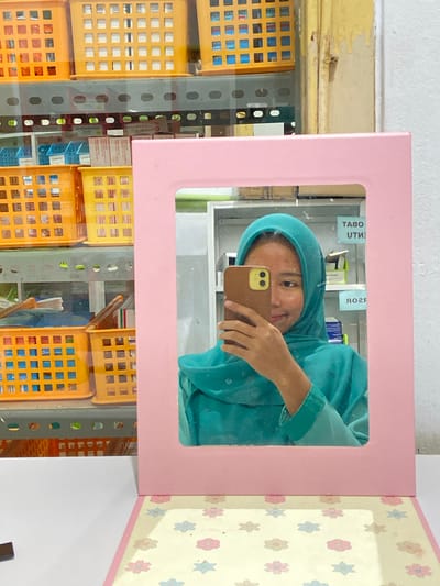 A young woman is taking a selfie in a mirror. She is wearing a teal hijab and a matching teal shirt. She is holding up a brown phone to take the picture. The mirror is framed in a pink border. The background of the mirror reflection shows shelves filled with various boxes of medicine and other items. The setting appears to be a pharmacy or a similar retail establishment. Outside the mirror, the scene continues with more shelves of colorful plastic baskets, likely used for organizing products. The baskets are yellow and orange, and some contain what appear to be small boxes or packets. A metal shelf unit is visible between the mirror and the baskets, suggesting a retail display. The overall impression is a brightly lit interior, possibly a storefront. The wallpaper behind the mirror has a pattern of small, colorful flowers. Some visible text in the reflection includes "UTM" and "ЯОЯ" on labels, as well as "Captopril" on a medicine box, indicating it's likely a pharmacy. The image is oriented vertically, suggesting it was taken with a phone in portrait mode.