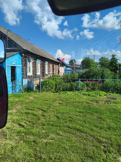 The image shows a rural scene in Il'mino, Russia, on a sunny day with a blue sky and white clouds. In the foreground, a patch of mowed grass is visible. To the left, the corner of a red vehicle is present. Behind the grass, a traditional wooden house with a dark roof and blue shutters stands. A bright blue wooden fence surrounds part of the property. Numerous green plants and bushes are growing in front of the house, with a cluster of pink flowers visible behind the fence. In the background, more trees and houses are seen, along with power lines.