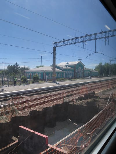 This is a medium-shot, slightly angled exterior view of the Chulym railway station in Chulym, Russia, taken on a clear day. The foreground shows construction work with orange netting fencing off a deep trench next to the railway tracks. The trench appears to be a work in progress, with exposed earth and some construction materials visible.
The middle ground features a light blue, single-story building with a white roof, identified as the railway station by a sign reading "ЧУЛЫМСКАЯ" (Chulymskaya) in Cyrillic lettering at the top of the main entrance. The building has arched windows and doors, and two white statues are placed outside on either side of the entrance. A paved platform runs in front of the station, and a person in a high-visibility vest is seen walking on the platform near a fence.
The background consists of trees and utility poles supporting overhead railway power lines. The sky is bright blue with a few faint white streaks, possibly reflections on the window through which the photo was taken. The overall scene depicts an active railway station undergoing some form of renovation or construction.