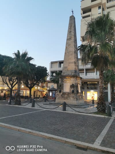 This is a medium-shot, eye-level, outdoor, daytime photograph taken in Follonica, Italy. The image depicts a stone obelisk with a star-shaped finial, situated in a paved town square. The obelisk is surrounded by a low stone wall and chains, with several palm trees flanking it. In the background, there are multi-story buildings with shops on the ground floor, some of which are illuminated. People are visible in the square, including shoppers and individuals sitting at outdoor tables. The sky is a clear, pale blue, indicating either early morning or late evening. The ground is paved with cobblestones. A watermark at the bottom left of the image reads "HUAWEI P30 Pro LEICA QUAD CAMERA".