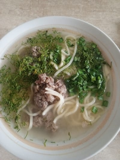 A close-up, overhead shot shows a bowl of soup filled with noodles, broth, minced meat, and fresh herbs. The bowl is white with a light beige rim and sits on a textured, light-colored surface, possibly a table.
The soup features thick, white noodles submerged in a clear, light-yellow broth. Scattered throughout the broth are pieces of seasoned minced meat, appearing as small, irregular clumps. A generous amount of chopped fresh dill and green onions is sprinkled on top. The dill is finely chopped and forms a feathery layer, while the green onions are cut into small, circular pieces, adding vibrant green accents.
The lighting suggests it's daytime, and the overall scene is casual and domestic. There are no people visible. The food appears to be a hearty, home-cooked meal, possibly a noodle soup with meat and fresh herbs, typical of Russian cuisine. The background is slightly blurred, focusing attention on the contents of the bowl. The setting appears to be an indoor environment, given the presence of a table and the diffused lighting. There is no visible text in the image. The presence of Buraly, Russia as location context suggests this meal might be prepared and consumed in that region.