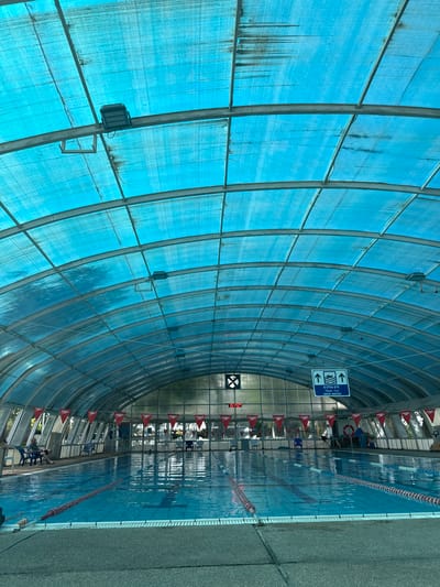 The image shows an indoor swimming pool in Beit Yitzhak-Sha'ar Hefer, Israel. The pool is covered by a translucent blue roof supported by a metal structure. The water is a bright blue with lane ropes visible. Along the sides of the pool, there are chairs and red Speedo triangular flags. A sign with arrows and the text "DEEP WATER" in English and Hebrew is hanging from the ceiling. Several people are visible, some sitting on chairs and one person appears to be in the water. The overall scene depicts a public swimming facility.