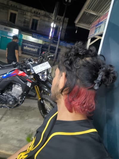 The image shows the back of a person with distinctive dyed red hair at the bottom, styled in buns with a clip. The person is wearing a black shirt with yellow accents on the sleeves and collar. A black and red motorcycle with gold accents is parked next to them. In the background, there are buildings that appear to be shops or businesses, with signs visible. The scene is lit by artificial lights, suggesting it is nighttime. One sign appears to be in Indonesian and has a blue and white design. Another sign, partially visible, has red and blue lettering. The setting seems to be an outdoor area in front of shops, possibly a street or a parking area. The overall impression is a casual, urban nighttime scene in Miruektaman, Indonesia.