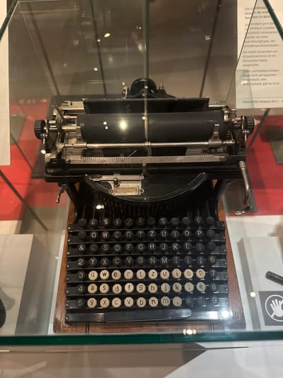 This media file shows a close-up, side-view of a vintage typewriter, likely displayed in a museum. The typewriter is black with silver metal accents and a wooden base. It features a full keyboard with circular keys, each bearing a letter, number, or symbol. Some keys are white, while others are black. The machine has a carriage with a roller and a ruler marking on its left side. Above the keyboard, a metal lever is visible.
In the background, through a glass enclosure, there are glimpses of exhibition displays. Text is visible on a white sign in the upper left, partially obscured by the typewriter. The text is in German and appears to be descriptive of the typewriter, mentioning "Speibmasc", "Revation im Büro", "Schreibmaschinen", and "Historisches Museum Bern".
The lighting suggests an indoor setting, possibly a museum exhibition, with bright reflections on the glass. No people are visible in the frame. The overall impression is of a historical artifact preserved and presented for public viewing. The German text indicates the location is the Historical Museum in Bern, Switzerland.