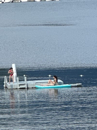 In the city of District of North Vancouver, Canada, two individuals are observed in the water on a bright, sunny day. One person, a woman, is lying down on a paddleboard. Another person, appearing to be a young girl, is standing on a floating dock. The water is calm, and there are several buoys visible in the distance. The scene suggests a recreational activity on a body of water, likely a lake or a bay.