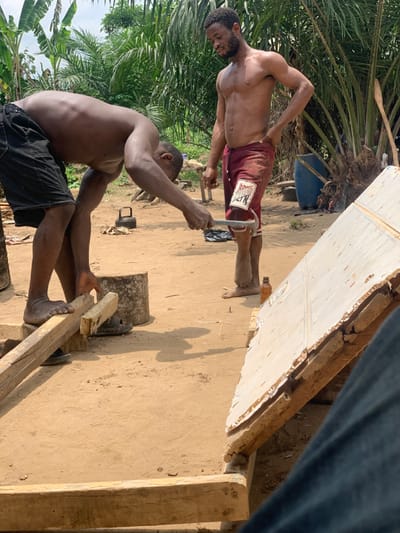 Two shirtless men are working on a wooden structure in Egbeda, Nigeria. One man is using a wrench to tighten a metal bracket. The second man is using a wooden plank to secure the structure. The men are both barefoot and wearing shorts. The structure is being built on a dirt floor and surrounded by vegetation. The location is near the coordinates 7.12 latitude and 5.362 longitude.