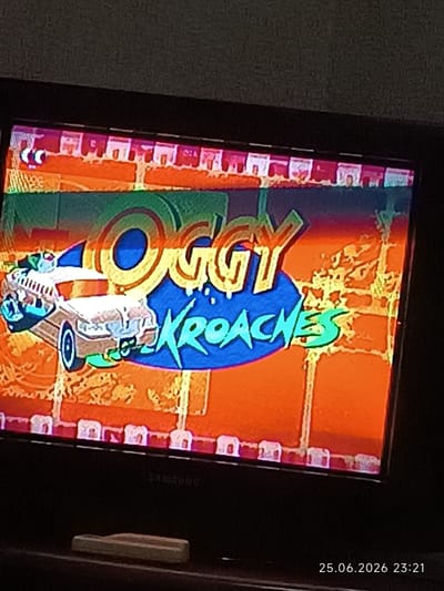 The image is a photograph of a television screen displaying the title screen of a cartoon. The screen shows a cartoon car, possibly a race car, with characters in it. The background is a stylized depiction of a brick wall with orange and green hues, and a blue circular logo. The title text reads "OGGY KROACHES" in a stylized font. The television is a flat-screen Samsung model. The bottom of the image shows a part of a remote control and a timestamp "25.06.2026 23:21" in the bottom right corner. The setting appears to be indoors, likely a living room or entertainment area, given the presence of the television and furniture. The image quality is somewhat blurry, suggesting it was taken with a mobile phone camera at an angle. The cartoon's title suggests an action or comedic theme, possibly a chase or a competition. The timestamp indicates the date and time the photo was taken, which is in the future. No specific weather or time of day details are discernible beyond what is implied by the indoor setting and the timestamp. The location context provided is Bolhrad, Ukraine, but there are no visual cues within the image to confirm this specific location.