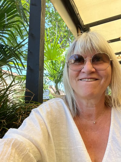 A close-up selfie depicts a woman smiling directly at the camera, seated outdoors under a cream-colored canopy supported by dark wooden beams. The woman has shoulder-length blonde hair with bangs, and wears large, round, gold-rimmed sunglasses with tinted lenses that reflect ambient structures and sky. She is dressed in a white, textured V-neck top and accessorized with a delicate gold chain necklace featuring a small, clear solitaire pendant. Her face shows some lines around her eyes and mouth as she smiles, revealing her upper front teeth which appear slightly uneven.

The background is rich with diverse green foliage, including prominent fan-shaped palm leaves and other dense greenery. A dark, vertically oriented wooden pillar, exhibiting a weathered texture, is visible through the leaves on the left side of the frame. Beyond the immediate foliage, glimpses of a light-colored building with a reddish-brown tiled roof are visible. On the right side of the background, partially obscured by greenery, a white directional sign is discernible, displaying fragments of text such as "rambert," "Service," "isme," and "40 Plag," which are consistent with the location of Saint-Just-Saint-Rambert, France. Overhead, clear, icicle-shaped decorative lights hang amidst the foliage. The scene is brightly illuminated by natural sunlight.