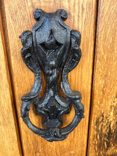 A close-up shot shows a black, ornate metal door knocker mounted on a light brown, wooden door. The knocker is composed of a decorative base and a ring-shaped handle. The wood has a visible grain pattern. The image was taken in Pulverbatch, United Kingdom.