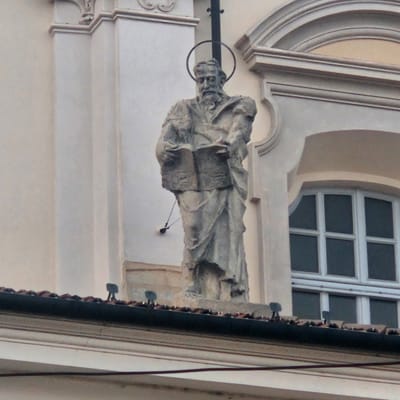 The image shows a stone statue of a bearded man holding a book, positioned on the roof of a building in Calcinate, Italy. The statue is weathered and appears to be ancient. A halo is visible behind the statue's head, suggesting it represents a religious figure. The building features classical architectural elements, including arched windows and decorative moldings. The sky is overcast, and the overall lighting suggests a cloudy day. The statue is centrally located in the frame, with the building's facade and roof details filling the background.