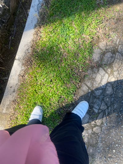 The image shows a person's legs and feet from a high-angle perspective, as if the photo was taken by the person themselves. The person is wearing white sneakers with velcro straps and black pants, with a hint of a pink garment (possibly a hijab or scarf) in the lower right corner. They are standing on a paved area next to a patch of green grass. The paved surface appears to be cracked and uneven, with small stones and dirt visible. The grass is vibrant green in some areas, with patches of dry, brown grass mixed in, suggesting it might be a naturalized or unmaintained area.
The scene is outdoors, with dappled sunlight casting shadows across the pavement and grass, indicating it is daytime. The presence of dry grass and the type of pavement suggest a warm climate, and the location context provided is Banjarbaru, Indonesia. There is no visible text in the image. The overall impression is casual and everyday, possibly taken while walking or taking a break outdoors.