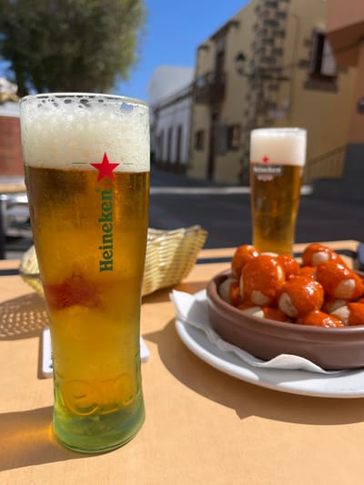 The image shows a wooden table with two glasses of beer, a small bowl of food, and a wicker basket to the left of the glasses. The glasses are filled with a light-colored beer. The glass on the left has a red star and "Heineken" written in green on the side. The glass on the right has a white logo on the side. The bowl contains orange-colored food with a sauce. The table and surrounding environment are lit by the sun. The scene is located in Agüimes, Spain, a small town on the island of Gran Canaria. The coordinates provided, 27.907 latitude and -15.447 longitude, are consistent with a location in Agüimes.