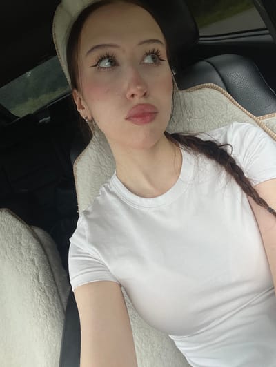 A young woman with light skin, long dark brown hair, and green eyes is sitting in the passenger seat of a car. She is wearing a white t-shirt and a light-colored headband. Her makeup includes winged eyeliner and pink blush. Her hair is in a braid that hangs over her shoulder. The car's interior is visible, including a black leather seat with a white fuzzy liner. The scene is not in Gavrilovka, Russia.