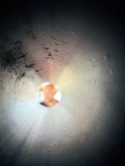 The image shows a close-up, macro view looking down a pipe or tube. In the center of the image, a blurred, spherical object with orange and white tones is visible, emitting a bright light that radiates outwards. The interior walls of the pipe are textured and appear to be a dark, metallic material, with some lighter, possibly condensation or dust, markings. The lighting is dim, creating a sense of depth and focus on the central object and its emitted light. No specific location in the city of Bašť, Czechia, can be identified from this abstract, close-up view.