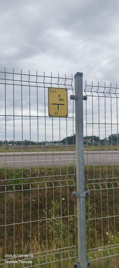 A yellow sign with black text and symbols is affixed to a metal fence. The sign reads "GAZ" at the top, followed by a circle with a diagonal line through it and the number "160". Below this are two horizontal rectangles with a vertical line connecting them in the middle, resembling a "T". At the bottom, the number "24" is displayed within a rectangle. The fence is gray and made of metal wires. In the background, there is a road with grassy areas and trees on either side. The sky is overcast. Text at the bottom left of the image indicates the date and time as "2026/07/09 06:16" and the location as "Stryków, Польша".