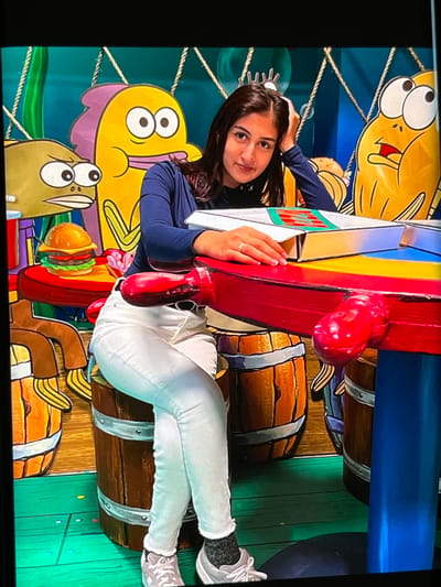 This media file features a young woman posing for a photograph in front of a colorful, cartoon-themed backdrop. The backdrop depicts stylized fish characters, ropes, and barrels, reminiscent of a playful, underwater or carnival setting. The woman is wearing a navy blue long-sleeved shirt, white pants, and athletic shoes. She has dark hair and is smiling at the camera, with one hand resting on a red, cartoon-like object that is part of the backdrop's display.
The scene appears to be indoors, likely in an entertainment venue or a themed exhibit, given the vibrant and artificial nature of the background. The lighting suggests it could be daytime, but the indoor setting makes it difficult to determine the time of day or weather conditions. The woman's expression conveys a sense of joy and engagement with the experience.
The backdrop itself is a notable detail, featuring exaggerated cartoon elements with bold outlines and bright colors. The presence of barrels suggests a potential theme related to pirates, treasure, or a nautical setting. The overall impression is one of fun and lightheartedness. No visible text is present in the image. The location context of Nijmegen, Netherlands, might imply this is a tourist attraction or a local event.