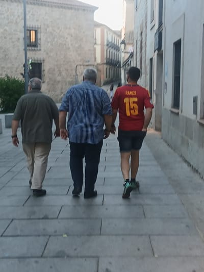The image shows three men walking away from the camera on a paved sidewalk in Ávila, Spain. The man on the left is wearing a short-sleeved olive green shirt and tan pants. The man in the middle is wearing a blue and white striped long-sleeved shirt and dark pants. The man on the right is wearing a red and yellow Spanish national team soccer jersey with "RAMOS" and the number "15" on the back, and dark shorts. He is also wearing colorful athletic socks and shoes. The buildings in the background are typical of Ávila's historic architecture, with stone facades and visible windows. The lighting suggests it is late afternoon or early evening.