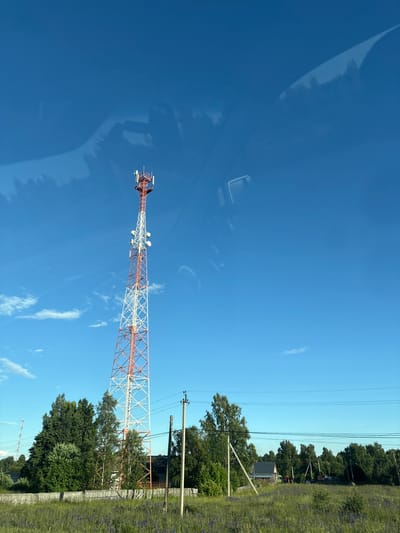 A telecommunications tower, painted in white and red, stands tall against a clear blue sky. The tower is the primary subject, with its intricate lattice structure reaching upwards. Around its peak, various antennas and equipment are visible. The image appears to be taken from a moving vehicle, as suggested by the reflections on a surface, likely a car window, which creates a blurred, abstract overlay on the sky.
To the right of the tower, a dense line of green trees forms a backdrop, partially obscuring some buildings and power lines. A grassy field, dotted with purple wildflowers, runs along the edge of the trees. Power lines stretch across the frame, supported by utility poles, indicating an inhabited or infrastructure-rich area. The lighting suggests it's daytime, with bright sunlight casting clear shadows. The overall scene is serene and rural, evoking a sense of open space and quietude. No people are visible in the frame, and no text is present.