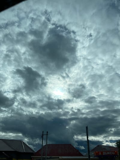 The image shows a cloudy sky above Semyonovskoye, Russia. The sun is partially visible through a break in the clouds. In the upper right, part of a building with a sign in Russian is visible. There are power lines cutting across the right side of the frame.