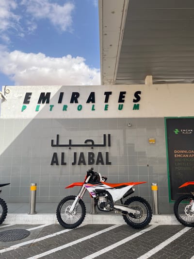 A vertically oriented shot shows a light gray building with "EMIRATES PETROLEUM" written vertically in English and a similar phrase in Arabic. Two orange and white KTM motorcycles are parked close to the building. A green advertisement is seen on the top right. The sky is visible in the bottom left corner, along with a few clouds. The ground is gray, striped with white lines.
