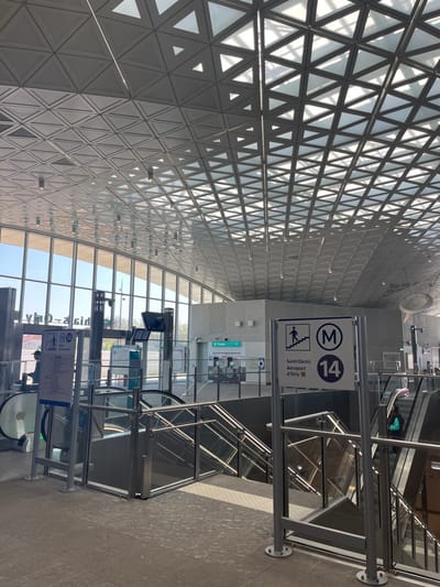 The image shows the interior of a subway station located at 48.747, 2.373 in Thiais, France. The scene features a large open space with a geometric ceiling made of triangular panels, and large windows. The space is primarily made of light grey tile. There are two escalators, one on the left and one on the right, which both lead down to a lower level. In the foreground, a person dressed in black is standing at the top of the escalator on the right. A sign, on the right wall, denotes the metro line as M14 and the destination as "Saint-Denis Aeroprt d'Orly." The sign is in French. There is another, smaller sign next to the main sign which depicts a person walking up a set of stairs and the words "Accès piétons d'Only," which means "Pedestrian Access Only" in French. A large window behind the sign shows a clear view of the outside. There are several other doors visible in the image, one of which is marked "Tokens." A person in a black jacket and a blue bag stands next to the escalator on the left. This person is looking toward the right. The background is blurred, showing other areas of the station. The lights on the ceiling are on, casting a bright glow throughout the space.