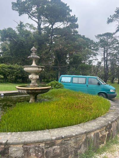 A blue van is parked on a driveway next to a stone fountain and lush green hedge. Tall trees and a cloudy sky form the backdrop. The fountain has multiple tiers and is surrounded by water. The driveway is paved with stone and bordered by a stone wall. The vegetation is dense and varies from manicured hedges to wild grass.