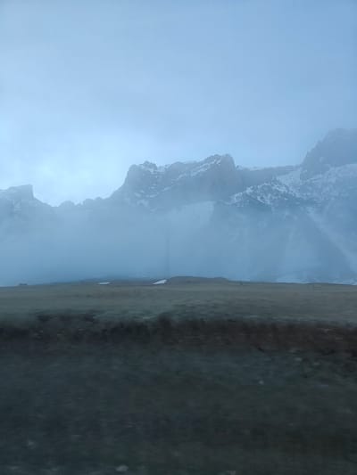 The image depicts a mountainous landscape shrouded in fog. The foreground features a dark, indistinct surface, possibly vegetation or ground cover. Above this, a flat, grassy area leads to a mountain range in the background. The mountains are partially obscured by heavy fog, with visible peaks covered in snow. The sky is overcast and light blue. The city of Kanobi, Georgia is not visible in this image.