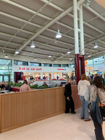 The image depicts an indoor scene within a shopping center, specifically located in Cannock Chase, United Kingdom. The environment is characterized by a high, corrugated metal ceiling with exposed beams and numerous pendant lights. Large white cylindrical pillars support the ceiling structure.
In the foreground, a wooden partition with vertical slats creates a defined area, possibly a seating or dining section. Beyond this partition, people are seated at tables, some engaged in conversation. A prominent red sign on the far wall reads "Exit to car park".
Further back, several retail storefronts are visible, including "Norton Canes Co-op" and a neon sign that says "ON - ULLY FAST FOOD". People are seen walking and browsing in these areas. The overall atmosphere suggests a busy public space. The people visible are dressed in casual attire.