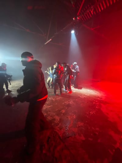 A group of people gathers under a spotlight, illuminated against a red-tinted background. The walls appear aged and textured, contrasting with the sharp focus of the light. One individual in a dark jacket with a red stripe across the back stands in the foreground. The scene is dimly lit, creating a sense of mystery. A light is attached to the ceiling to the left.