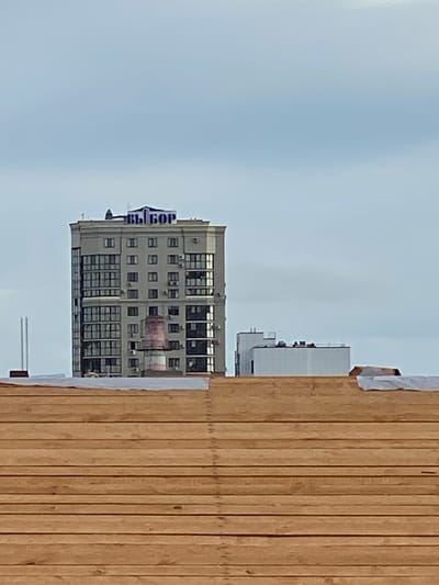 The image shows a multi-story residential building with a sign that reads "ВЫБОР" in blue letters at the top. The building is light-colored with numerous windows. To the right of the main building, there is a smaller, white building with a flat roof. In the foreground, there is a structure made of horizontal wooden planks, partially covered with white sheeting. The sky is overcast and appears to be a light blue-gray color. There are no people visible in the image, and no specific location information, such as Yokohama, Japan, is present within the image itself. The image is a photograph taken from a distance, looking up at the buildings.