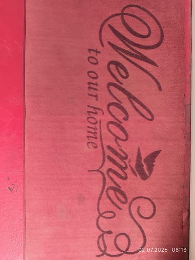 The image displays a close-up view of a pink welcome mat with dark red lettering. The mat is oriented vertically, with the text "Welcome to our home" written in a cursive font. A stylized bird or butterfly graphic is also present on the mat, positioned near the "e" of "Welcome." The texture of the mat is visible, appearing to be a rough, looped material. To the left of the mat is a bright pink, smooth surface that appears to be a wall or a similar flat surface. The right edge of the image shows a sliver of a white vertical object. A timestamp "02.07.2026 08:13" is present in the bottom right corner of the image. The scene is indoors or an entryway and is devoid of people or any other discernible activity. The location is specified as Kattasu, Uzbekistan.